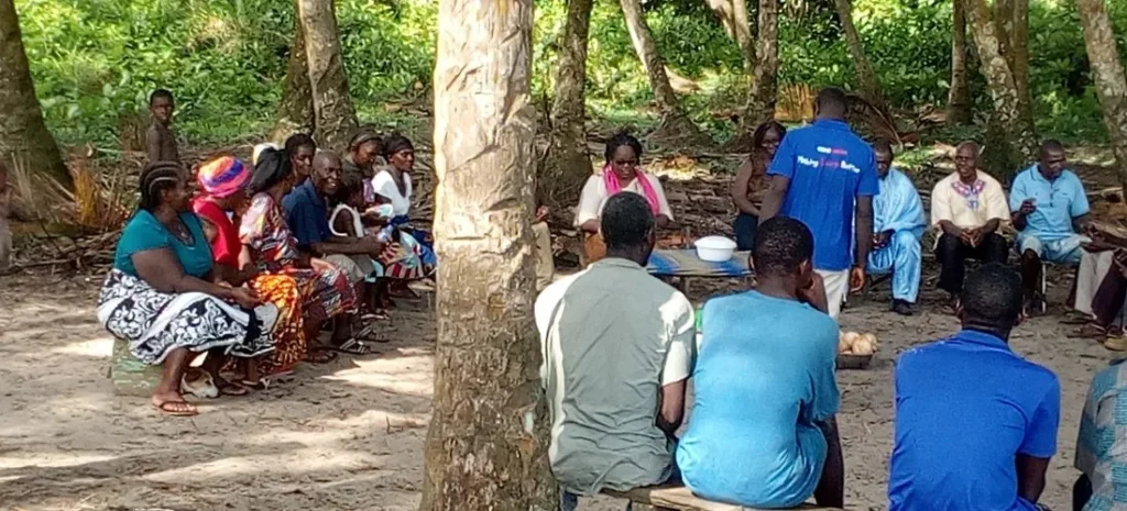 Community members of Grandcess seated together under palm trees during a village meeting